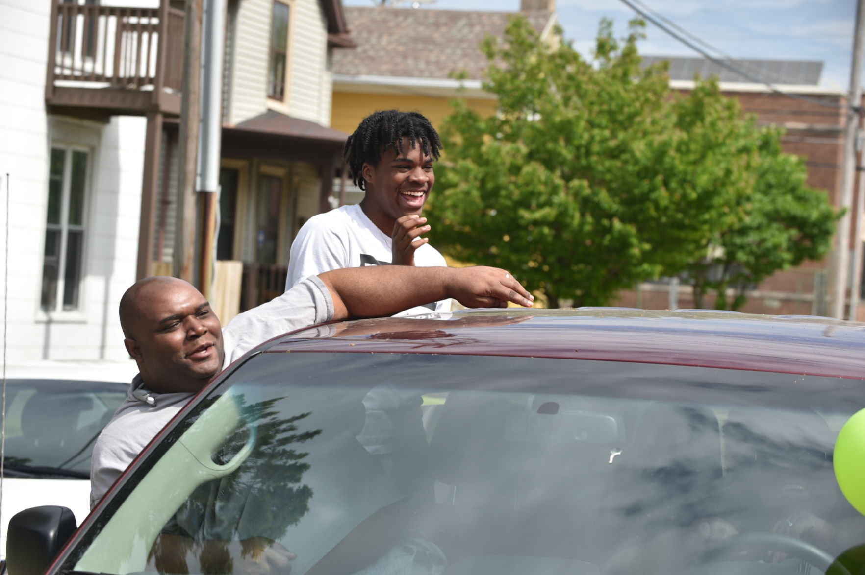 Two men laugh and hang out of a van while cheering on Bishop Kirby